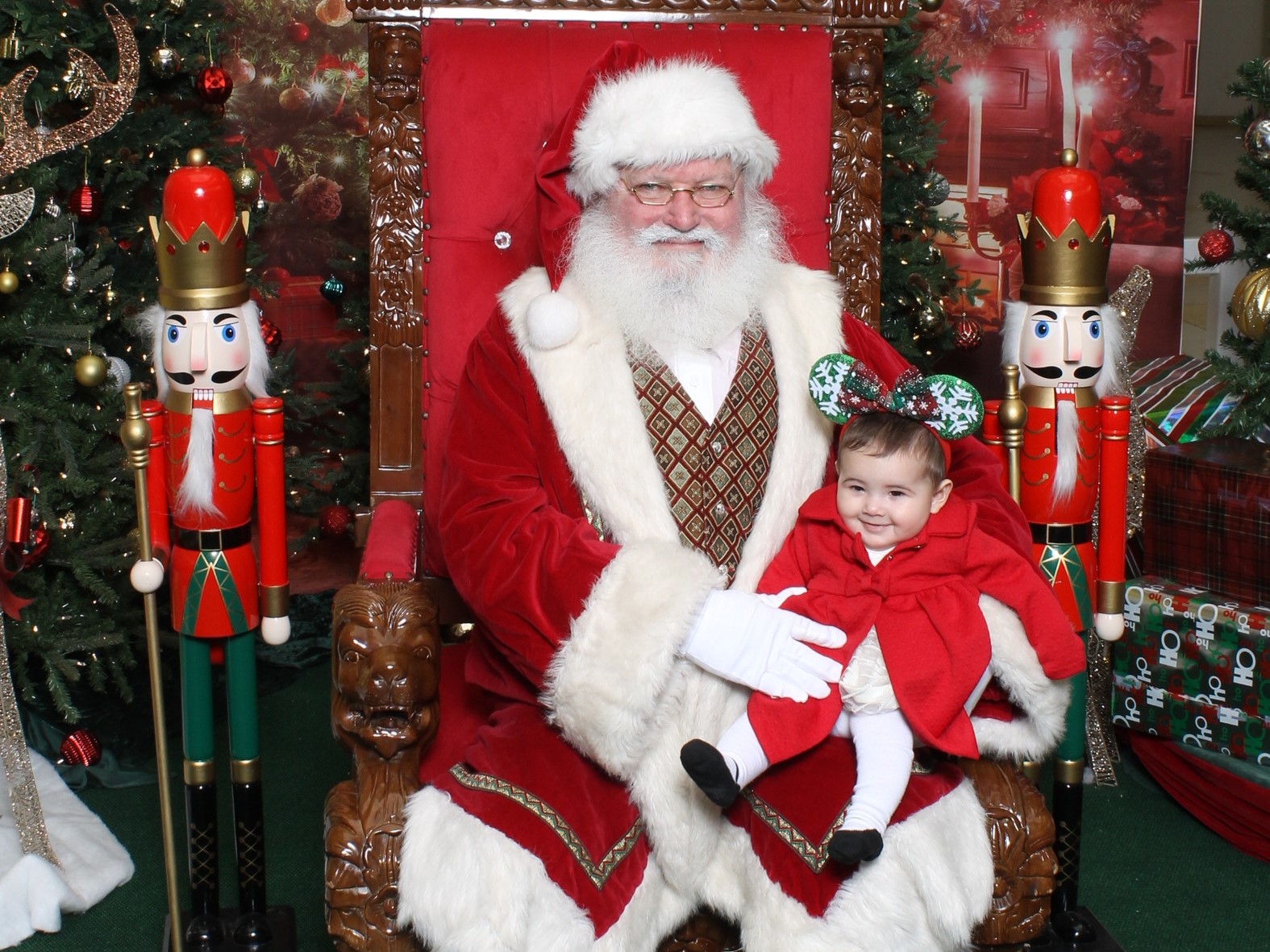 Santa sits on a grand throne, holding a child dressed in a festive red outfit, flanked by nutcracker figurines amidst a Christmas backdrop.